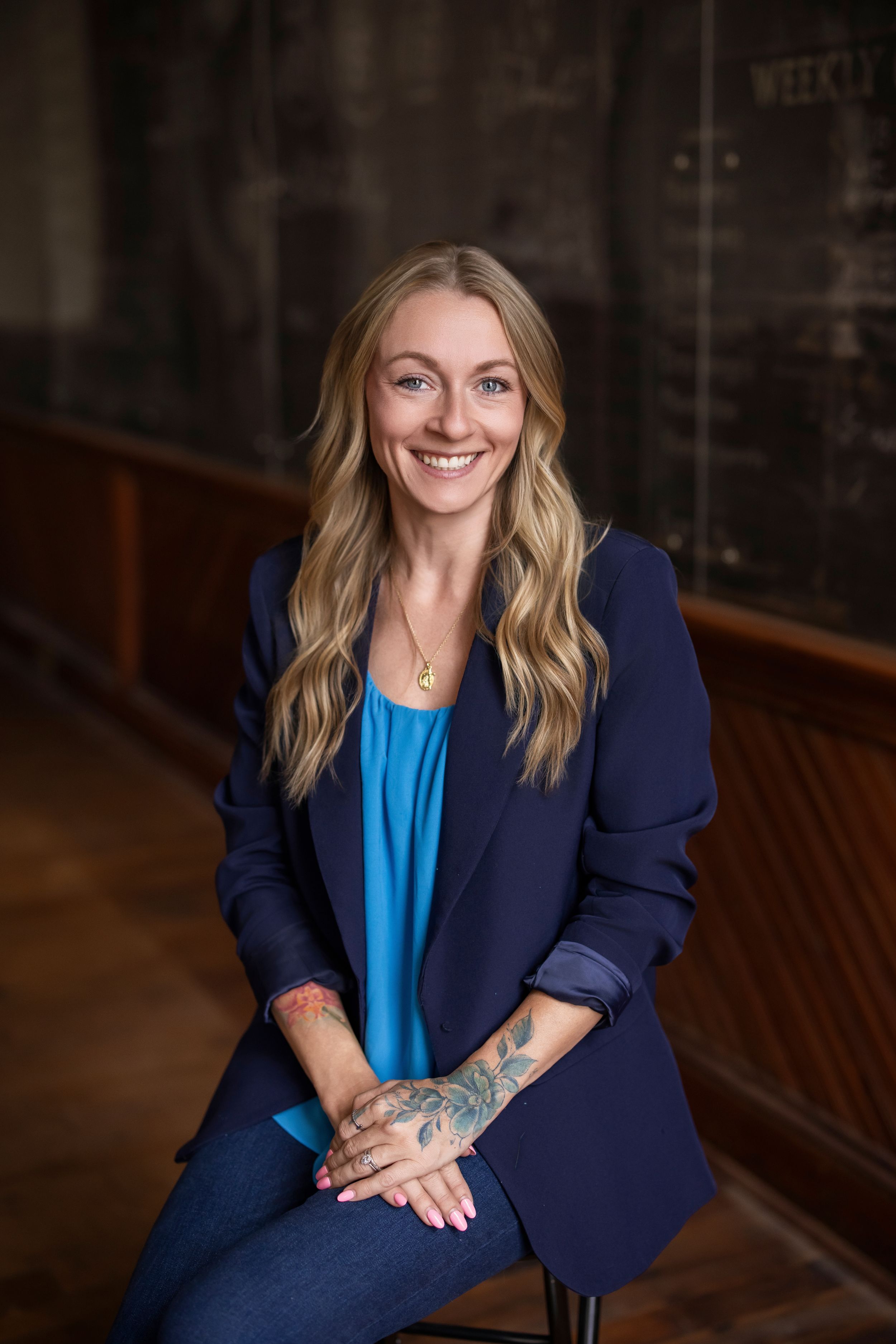 Smiling blonde woman with tattoos in navy blazer and blue top seated in a professional office setting.