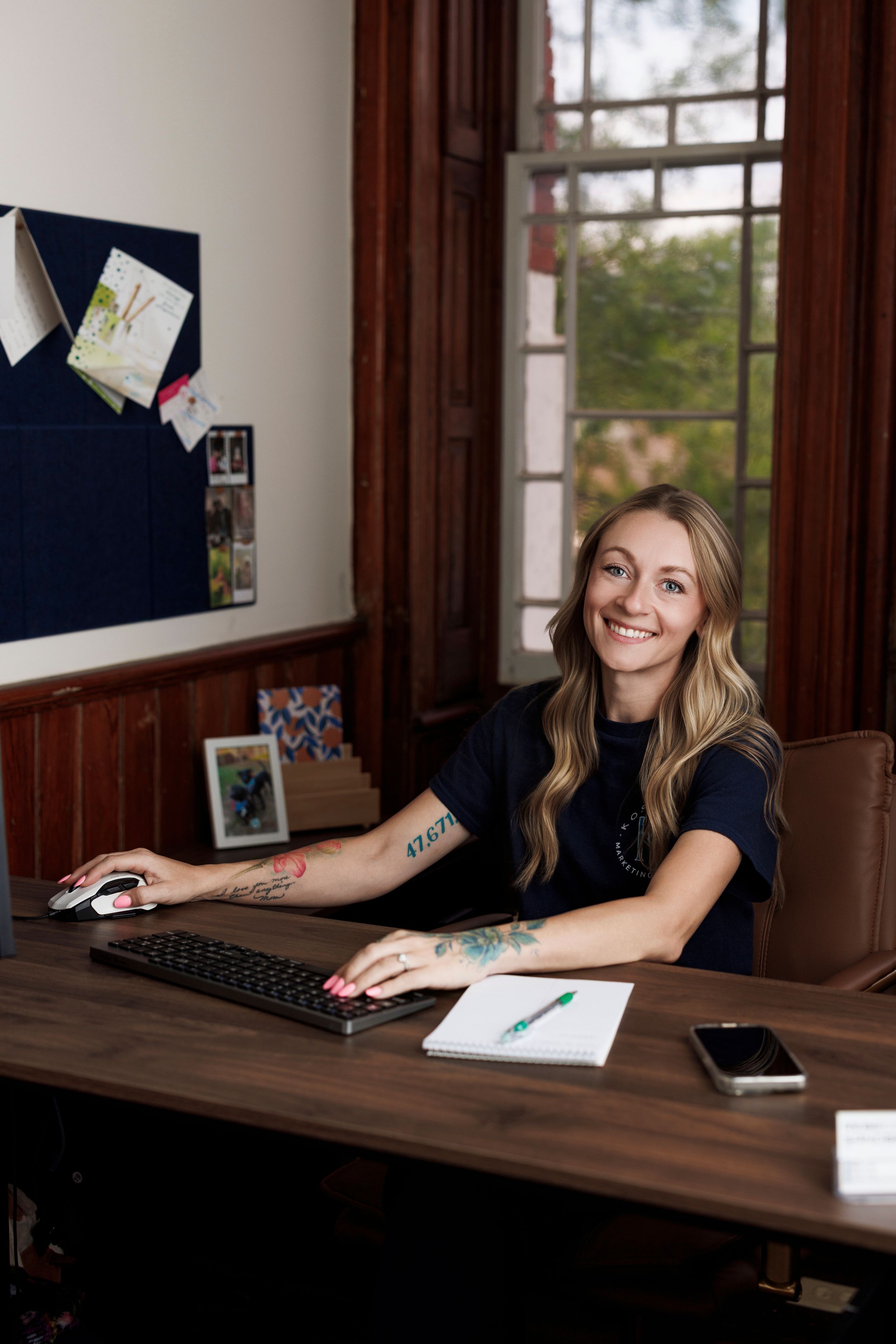 Smiling woman with tattoos sitting at a wooden desk using a computer keyboard and mouse in a cozy office.