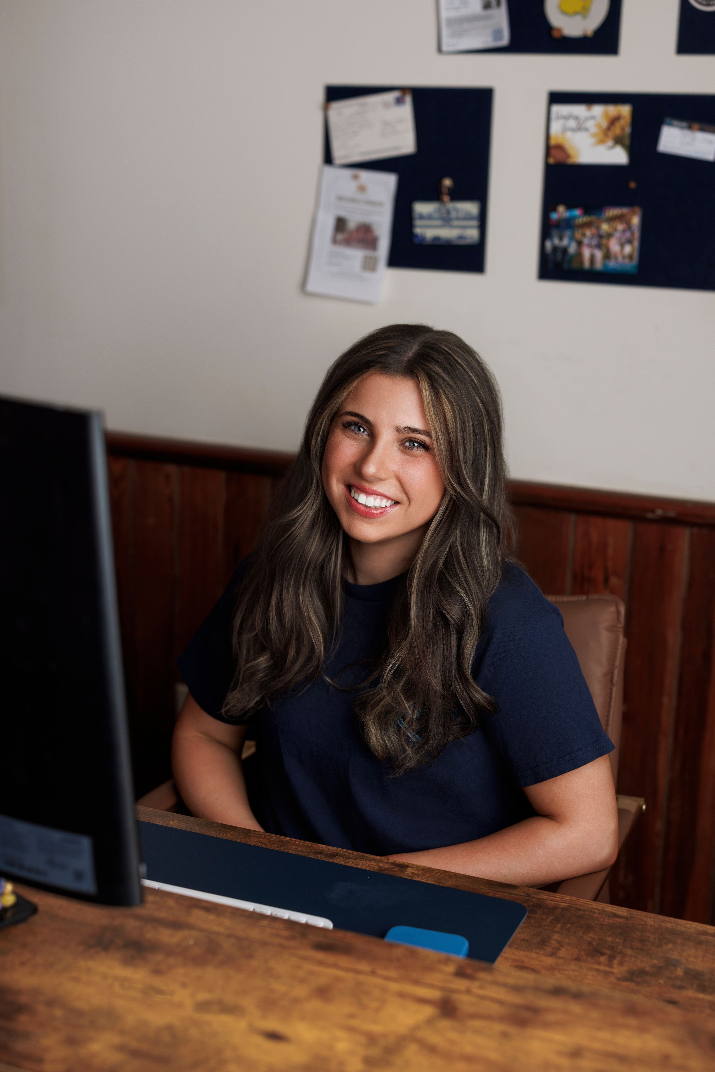 Smiling young woman with long dark hair sitting at a wooden desk with a computer in a cozy office.