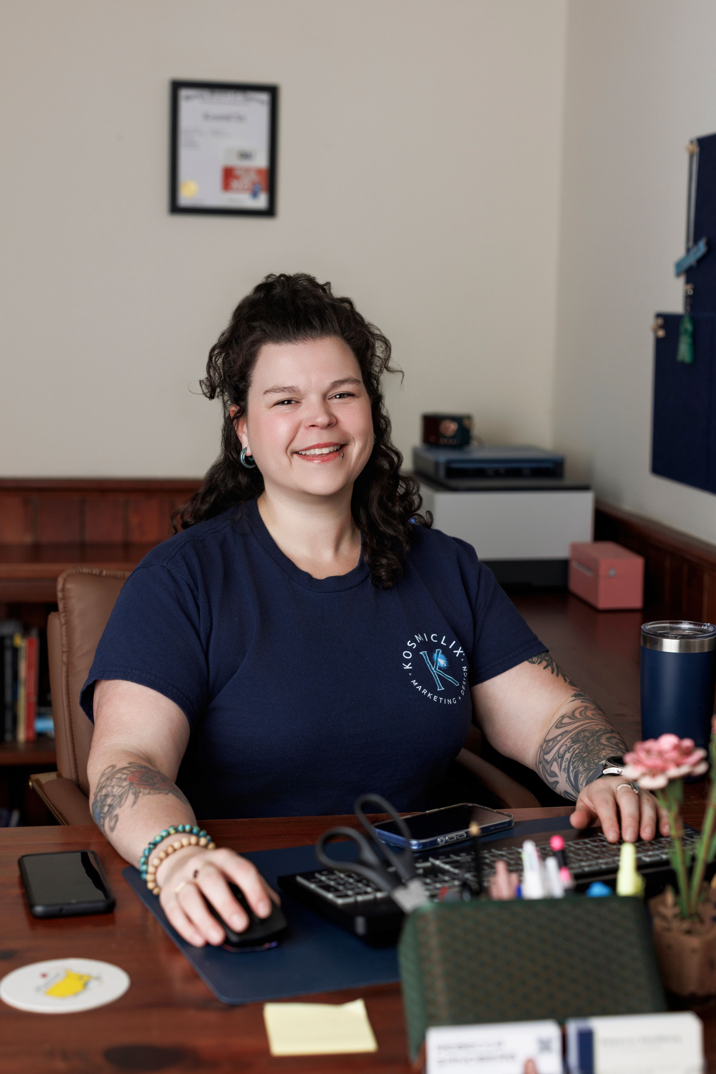 Tattooed woman with curly hair smiling at a desk with office supplies in a bright workspace.