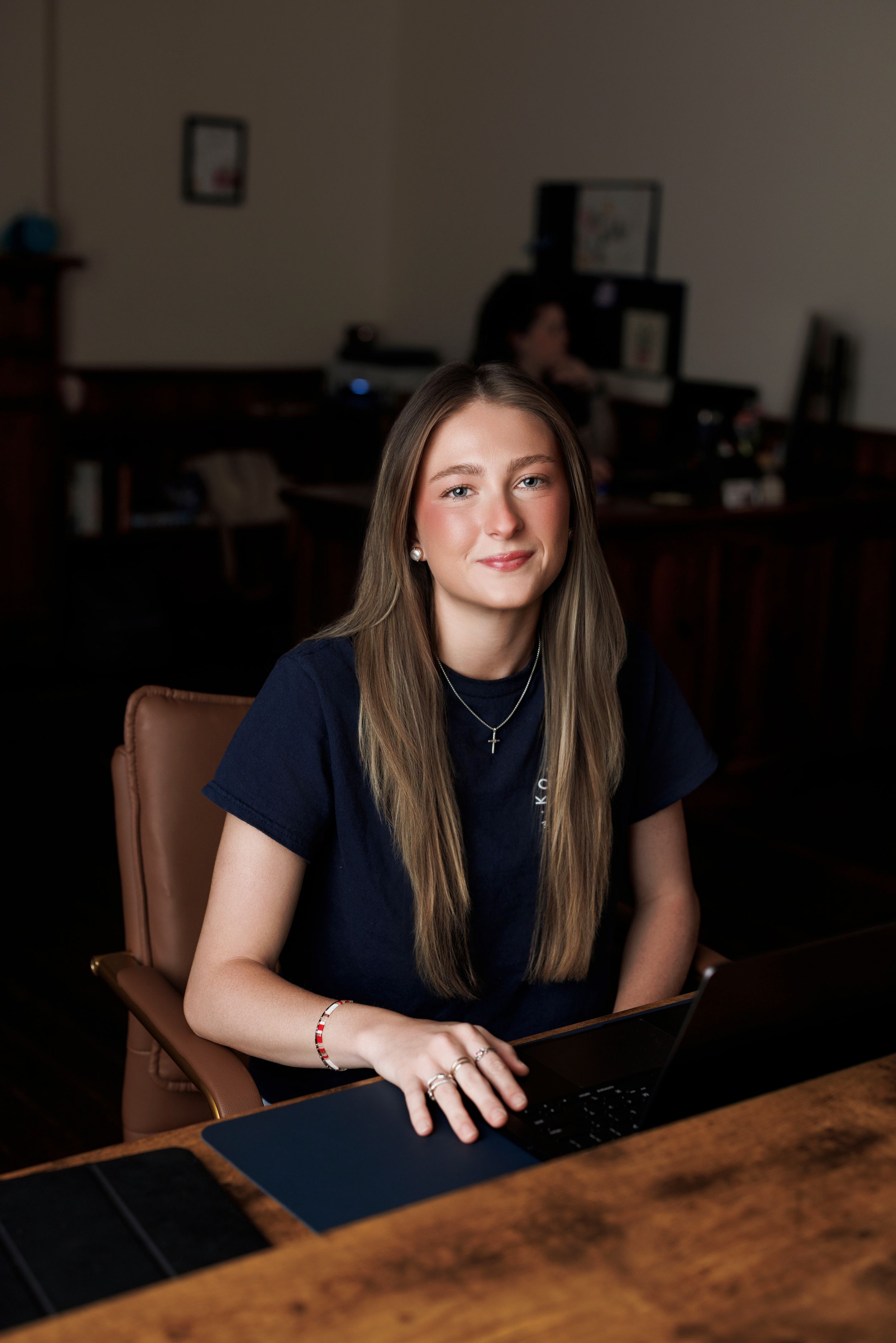 Young woman with long hair in navy top working on laptop at wooden desk in office environment.