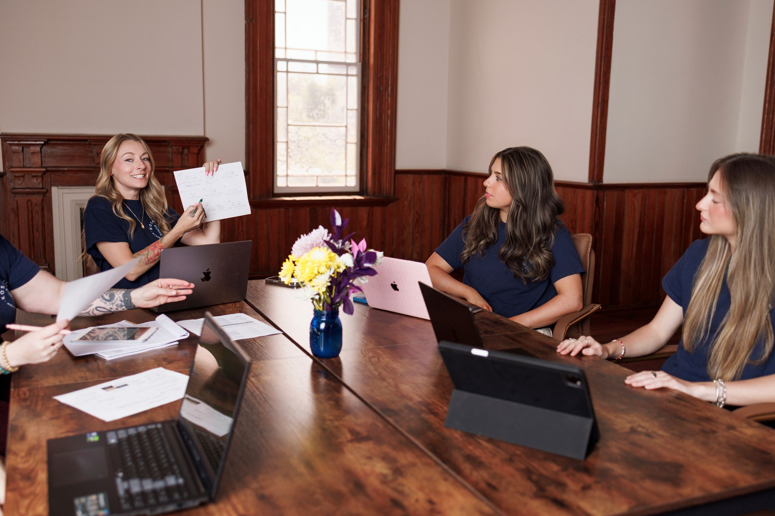Women in navy shirts collaborate around a wooden conference table with laptops, documents, and a floral centerpiece.
