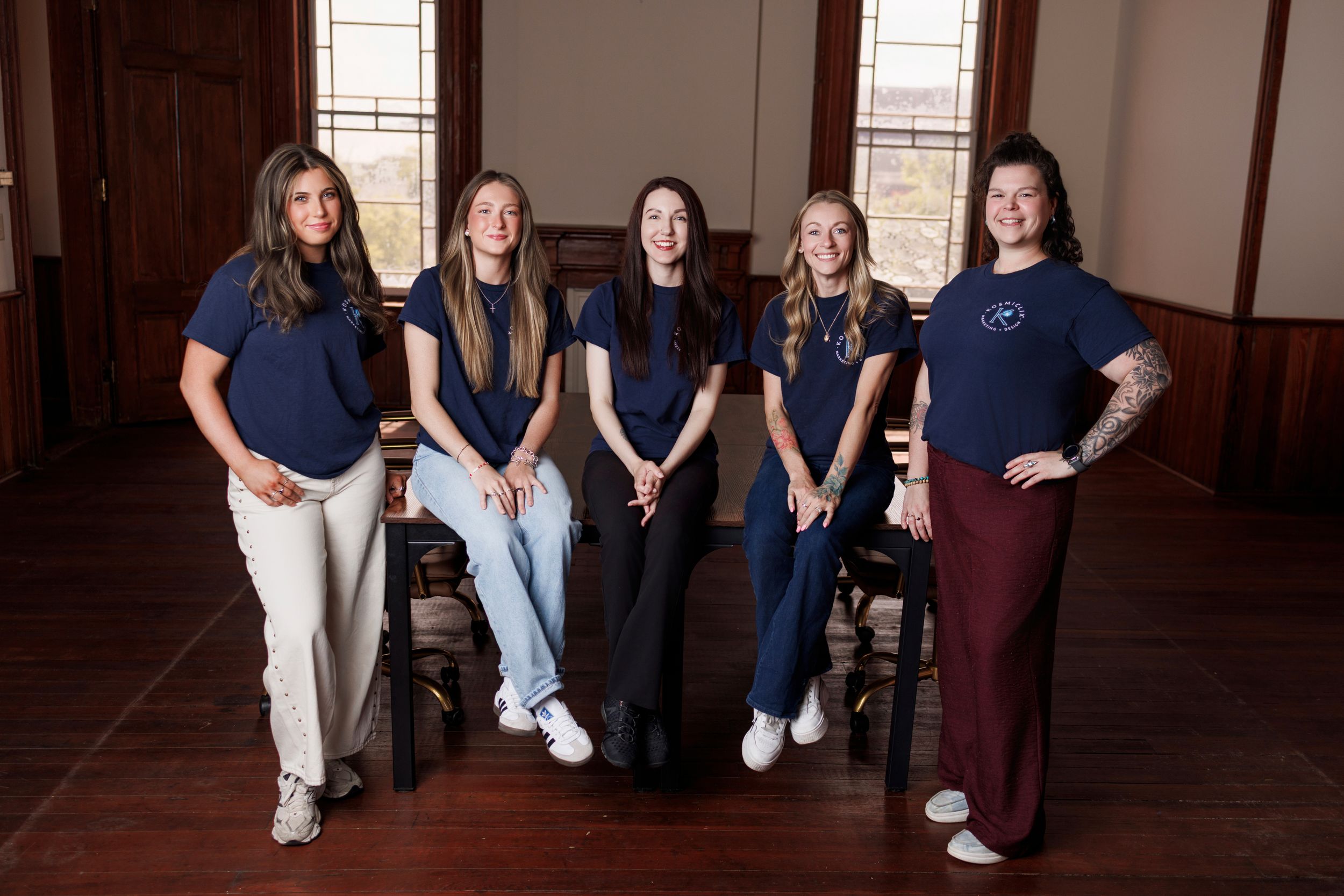 Five women in matching navy blue t-shirts pose together smiling in a warmly lit wooden room.
