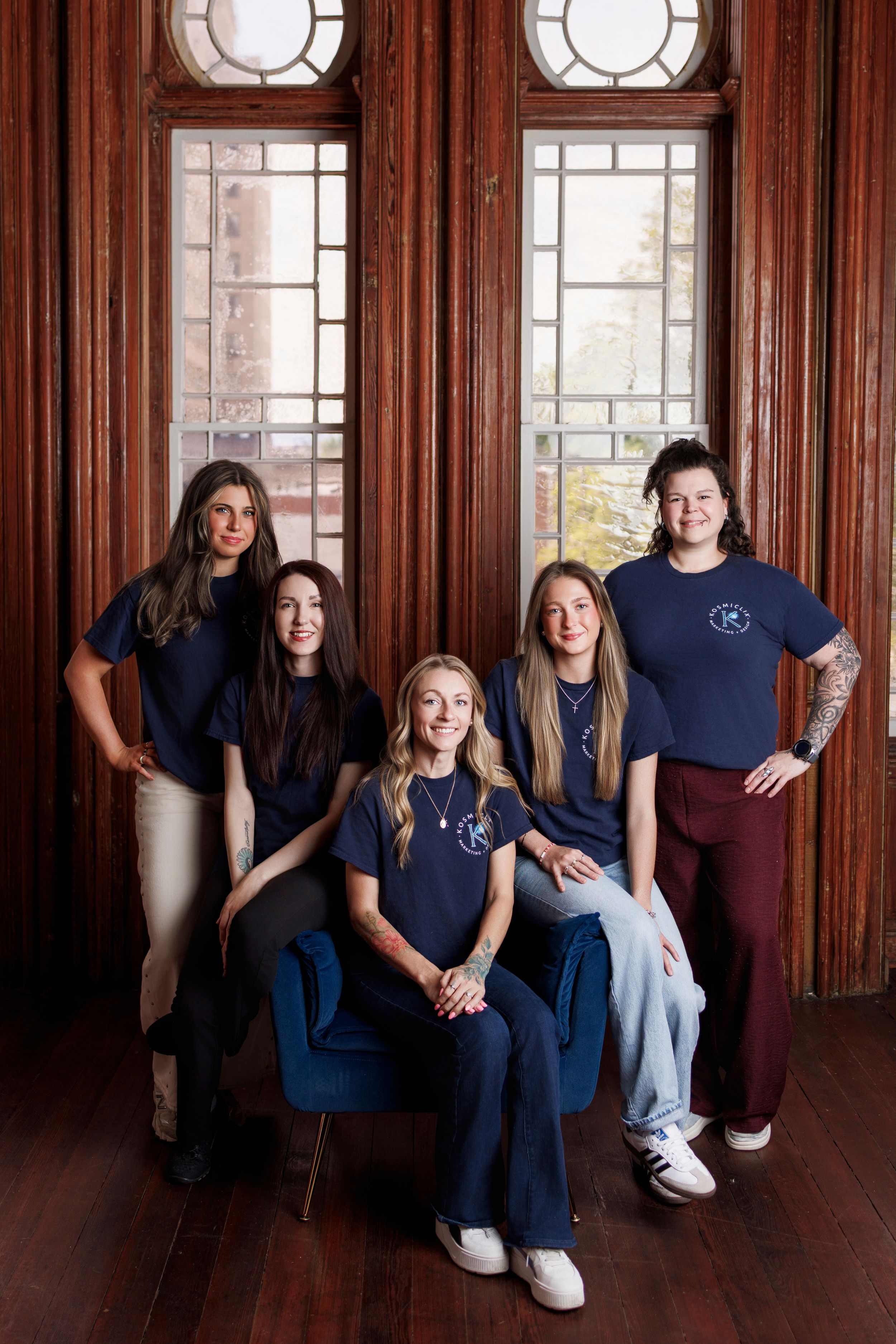 Group of five women in matching navy blue t-shirts posing together in a wood-paneled room with large windows.