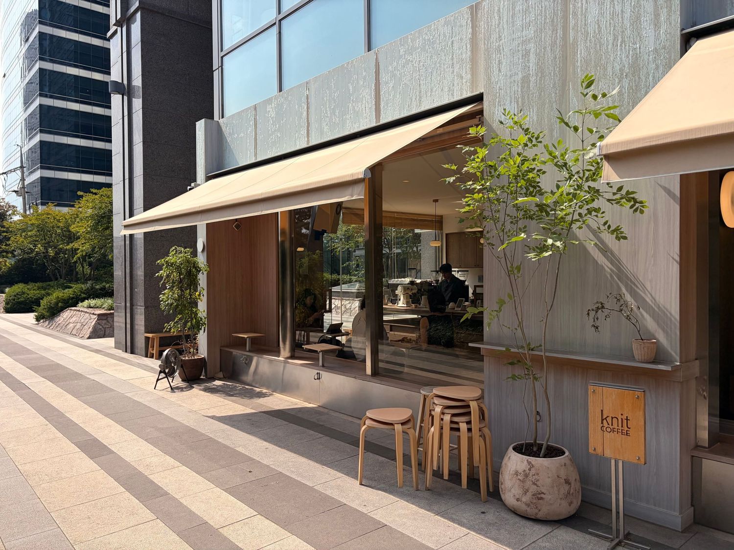 Modern Japanese cafe exterior with wooden stools, potted trees, and retractable awning on a sunny urban sidewalk.