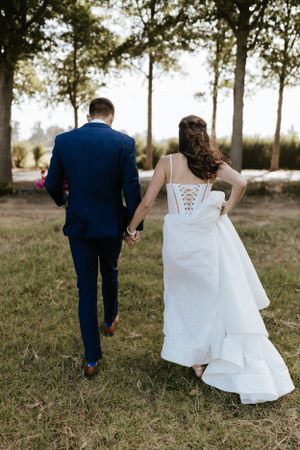 Back view of newlyweds walking hand in hand, showing bride's lace-up dress detail and groom's navy suit.