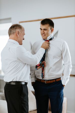 Two people in formal attire helping with tie adjustment during wedding preparations.