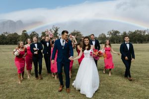 Wedding party celebrates under a rainbow in an open field with bridesmaids in coral dresses and groomsmen in dark suits.