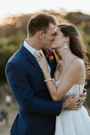 Romantic sunset wedding portrait of couple embracing and kissing in formal attire.