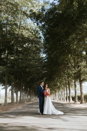 Newlyweds pose in a dreamy tree-lined path holding a coral bouquet.