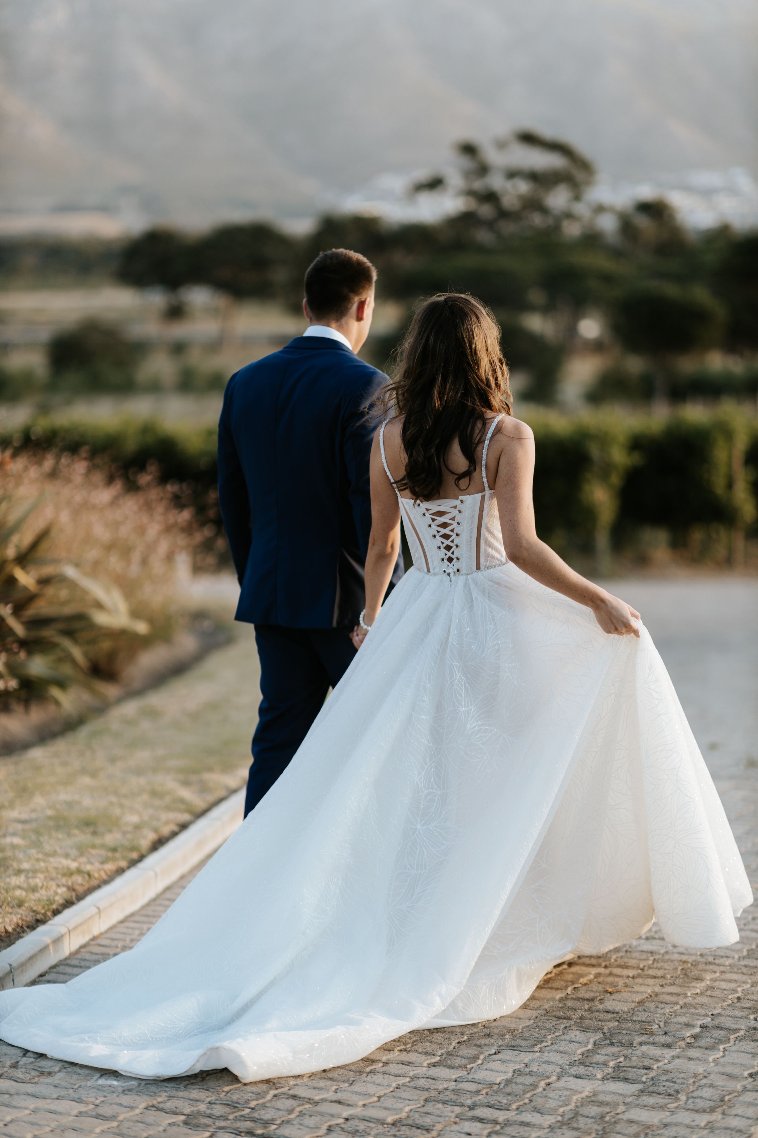 Newly married couple walking down path at sunset, bride wearing corset back white gown and groom in navy suit.