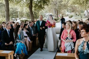A wedding processional at an outdoor ceremony with guests seated on wooden benches.