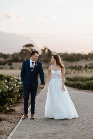 Couple walking hand in hand on pathway surrounded by vineyards at golden hour.