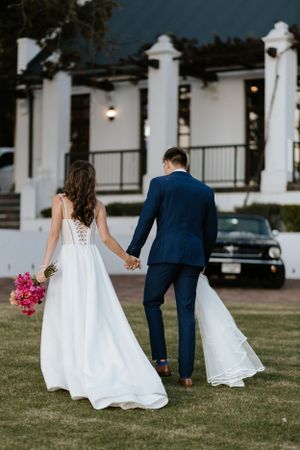 Couple walking hand in hand with pink bouquet towards white columned building.