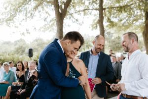 An emotional moment during an outdoor wedding ceremony with guests looking on beneath trees.