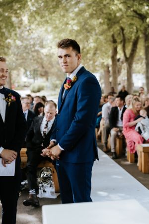 Groom in navy suit standing at outdoor ceremony with wooden benches and trees.
