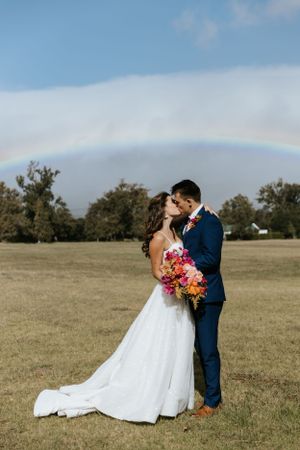 A romantic wedding portrait under a rainbow with a vibrant pink and orange bouquet.