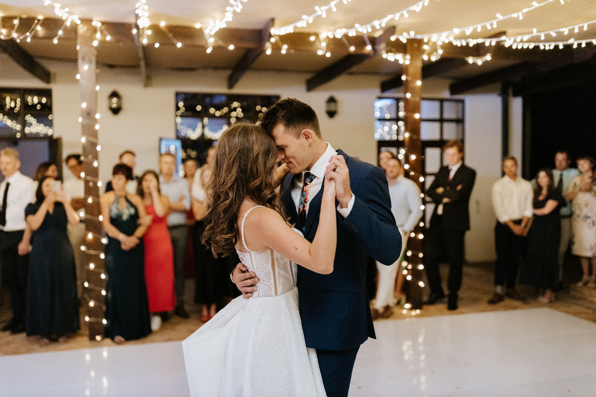 Newlyweds dancing together in a venue decorated with twinkling lights while guests look on.