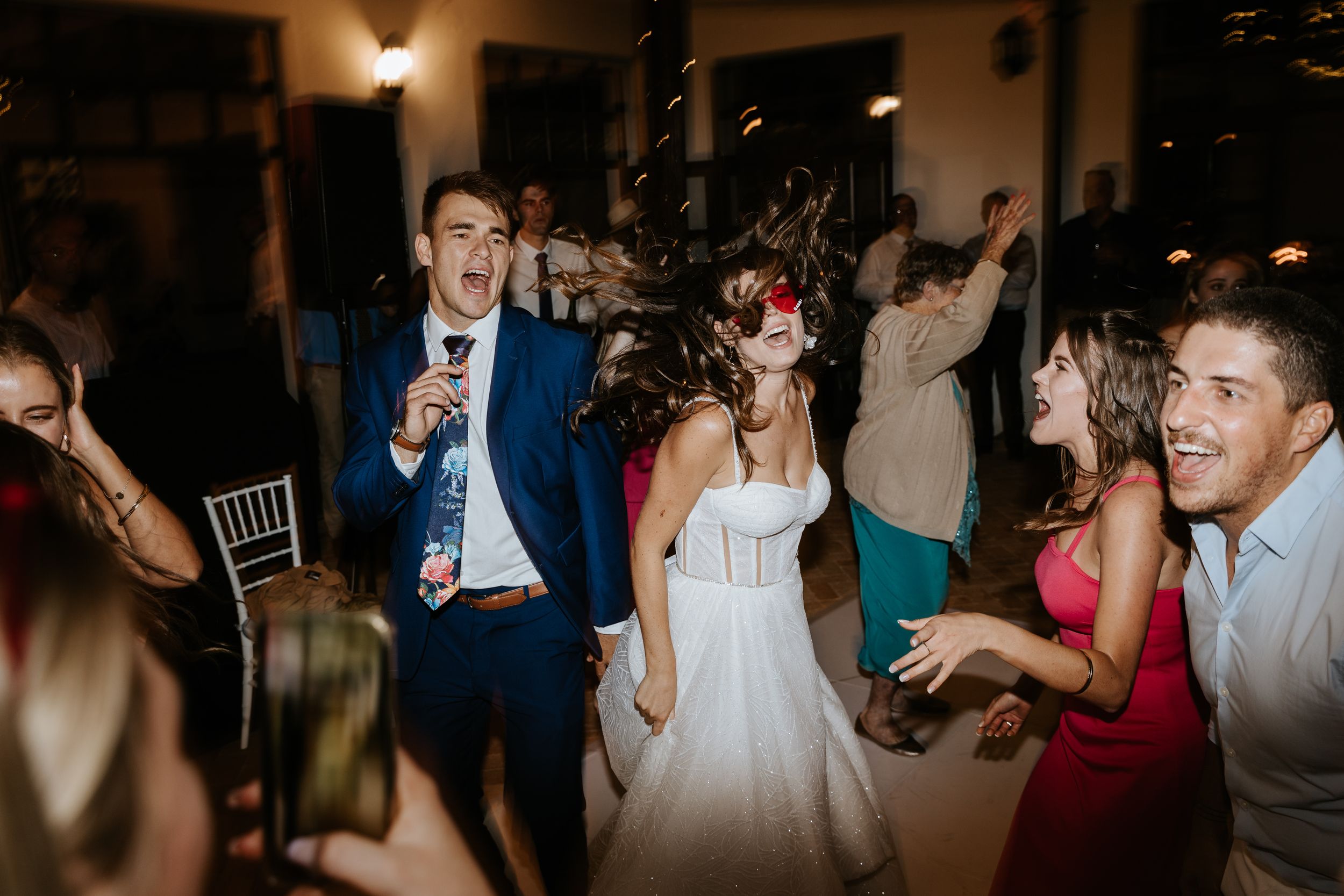Wedding party dancing energetically at reception with bride in white dress and guests in colorful attire.