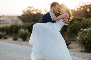 Groom lifts bride in flowing white dress during sunset portrait session.