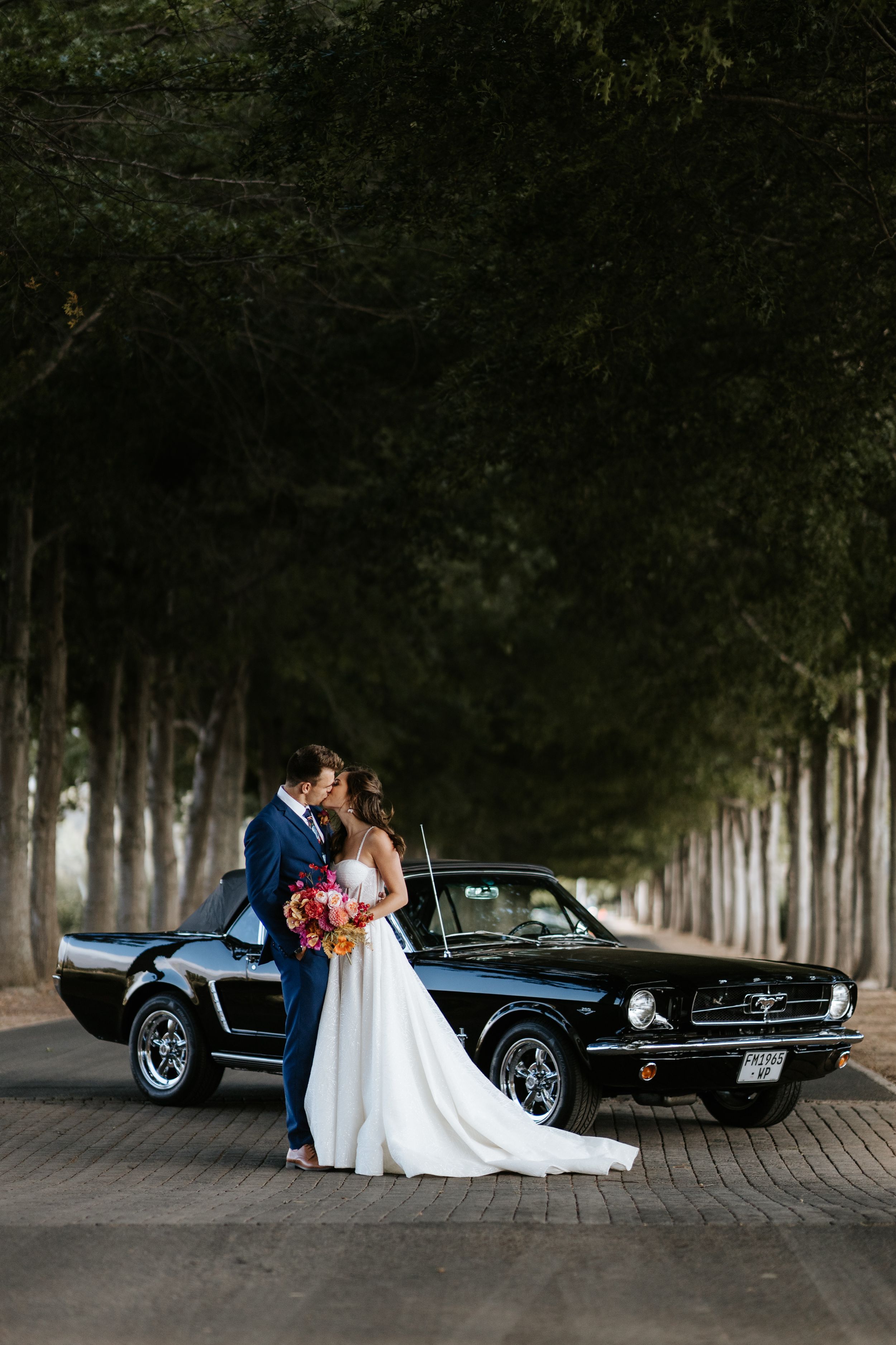 Elegant wedding couple poses with classic black Mustang on a tree-lined road.