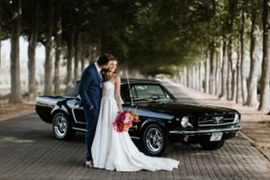 Newlyweds share an intimate moment beside vintage black Mustang on brick-paved avenue.