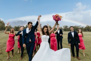 Joyful wedding party jumps and poses together in a field under a rainbow with bright coral bridesmaid dresses.