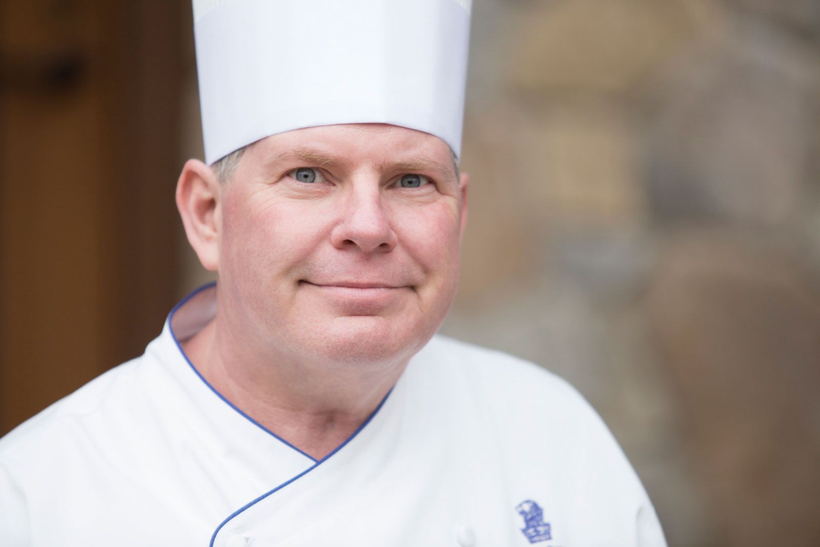 Professional chef in white uniform and tall toque hat smiling warmly at camera outdoors.