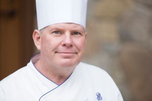Professional chef in white uniform and tall toque hat smiling warmly at camera outdoors.