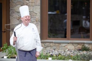 Smiling chef in white uniform holding garden rake outside stone building with herb garden.