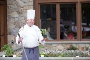 Chef in white toque holding garden hoe and small potted plant outside stone restaurant.