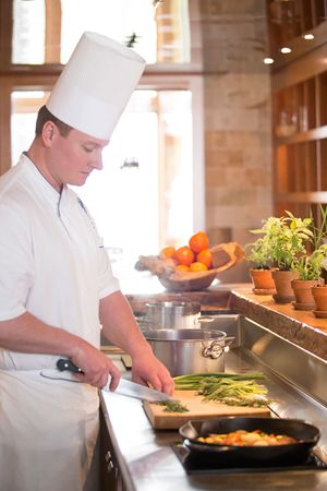 Chef in white uniform chopping fresh green asparagus on cutting board in professional kitchen.