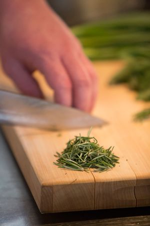 Chef mincing fresh rosemary herbs with knife on wooden cutting board.
