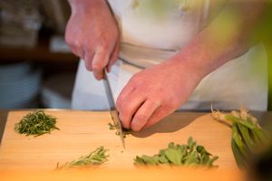 Chef chopping fresh rosemary and sage herbs on wooden cutting board.