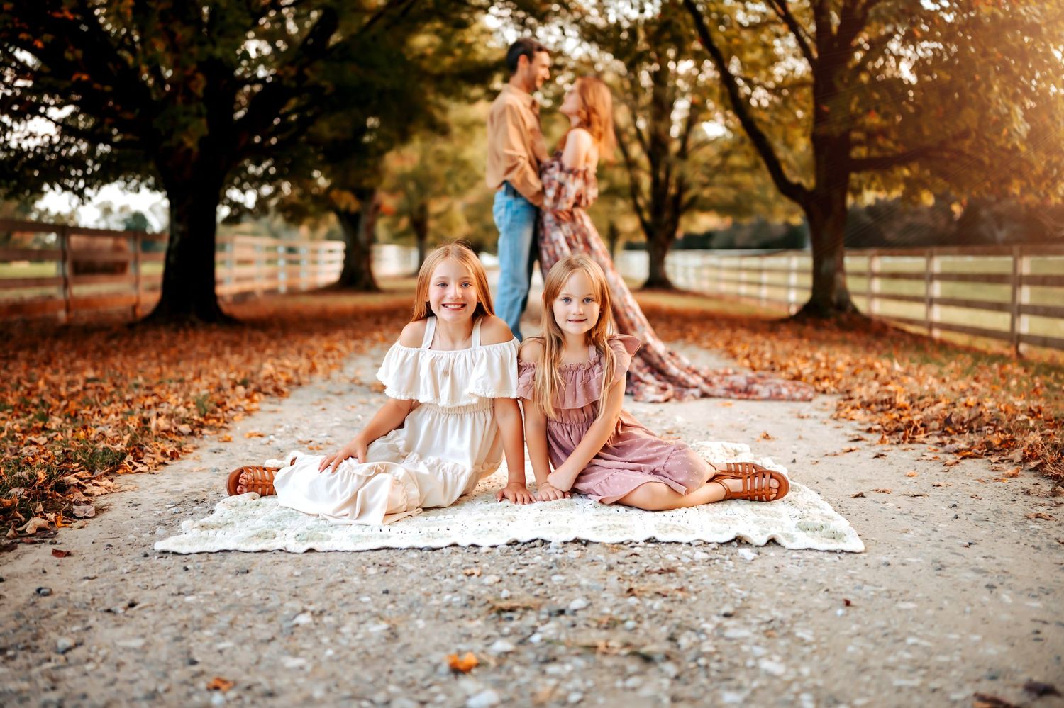 Two blonde girls sit on a lace blanket outdoors in fall, parents embracing behind them among golden leaves.