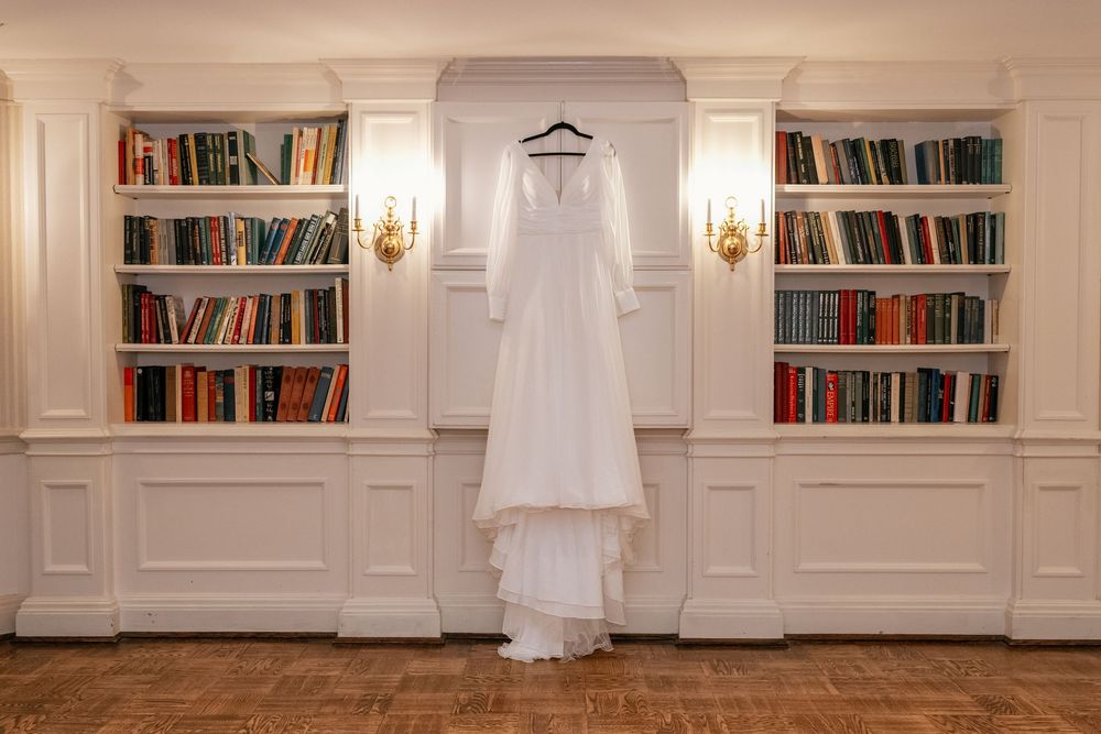 White wedding dress hanging on a hanger between two white bookshelves with colorful books and gold wall sconces.
