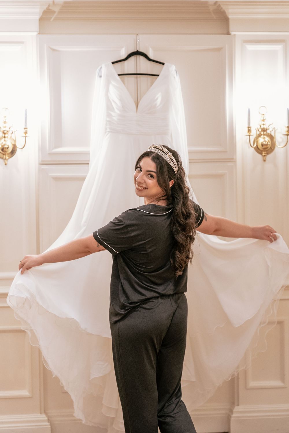 Bride in dark pajamas holding up her white wedding gown in an elegant room before getting dressed.