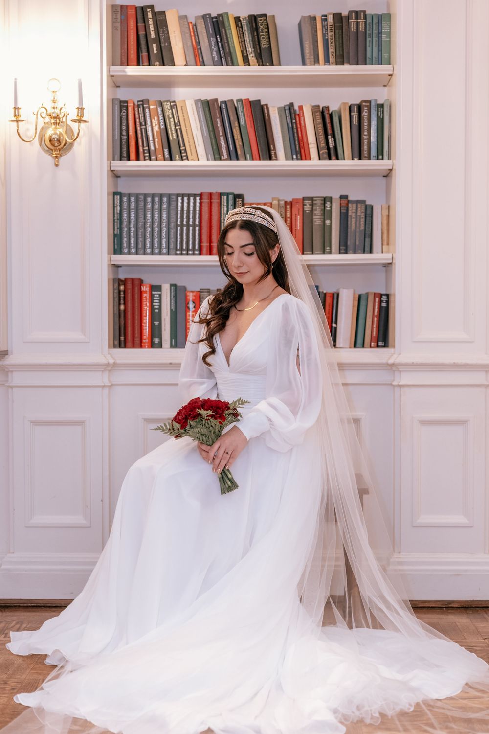 Elegant bride in white gown with veil holding red rose bouquet seated by a bookshelf.