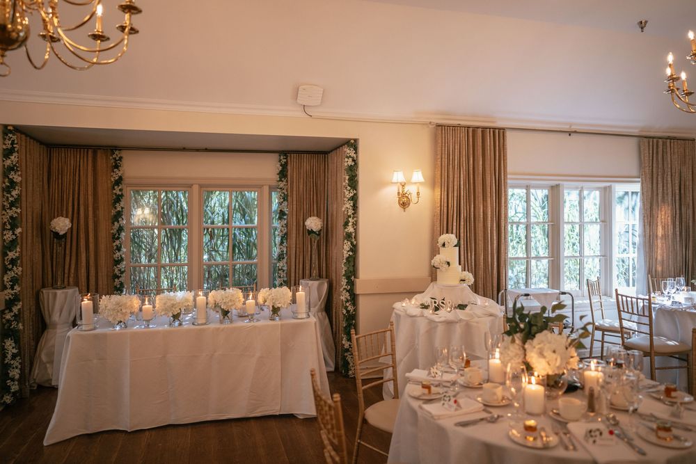 Elegant wedding reception room with white tablecloths, candles, tiered cake, and gold chairs under warm chandelier lighting.