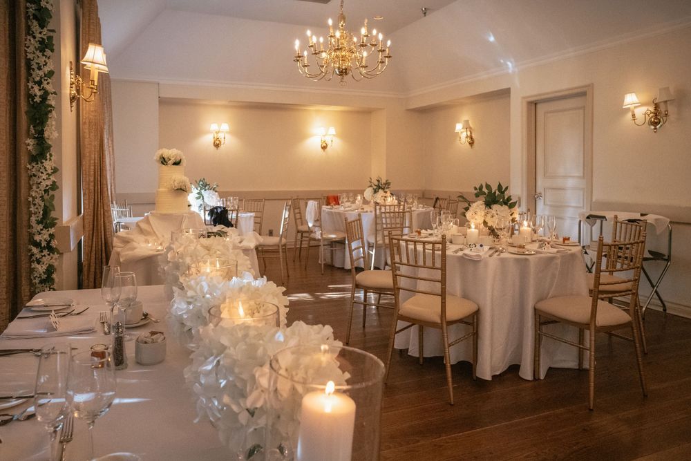 Romantic wedding banquet hall featuring white floral centerpieces, candles, gold chiavari chairs, and a crystal chandelier.