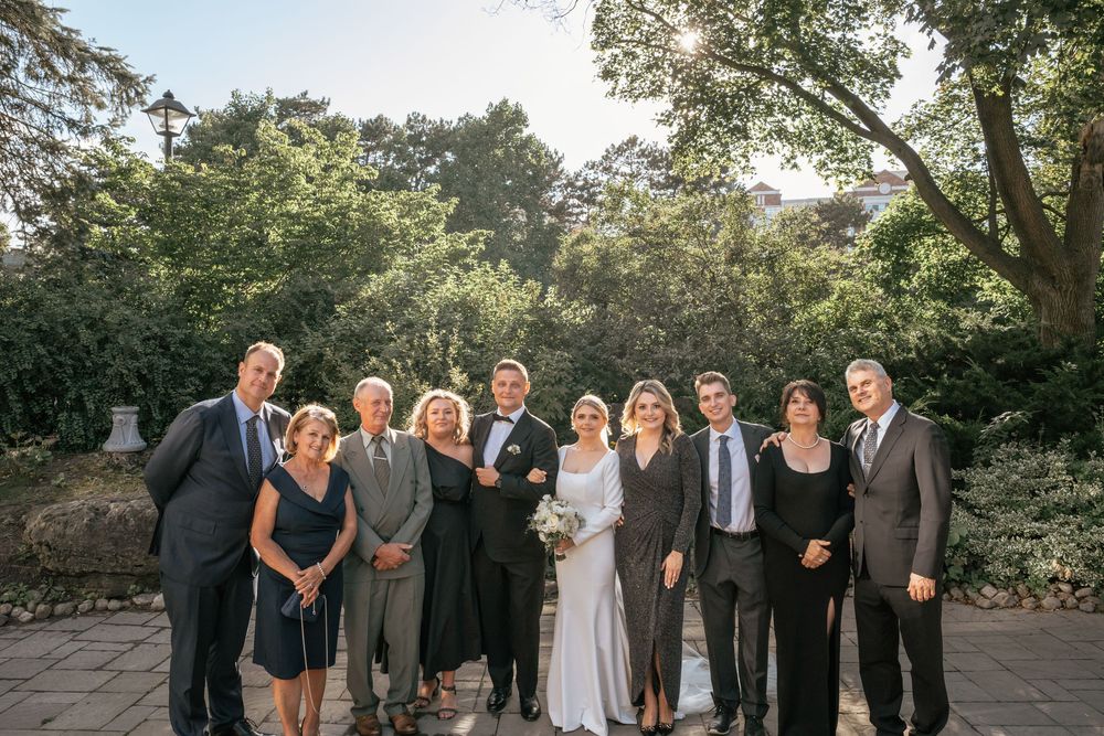Wedding party group photo with bride and groom surrounded by family in elegant attire outdoors.
