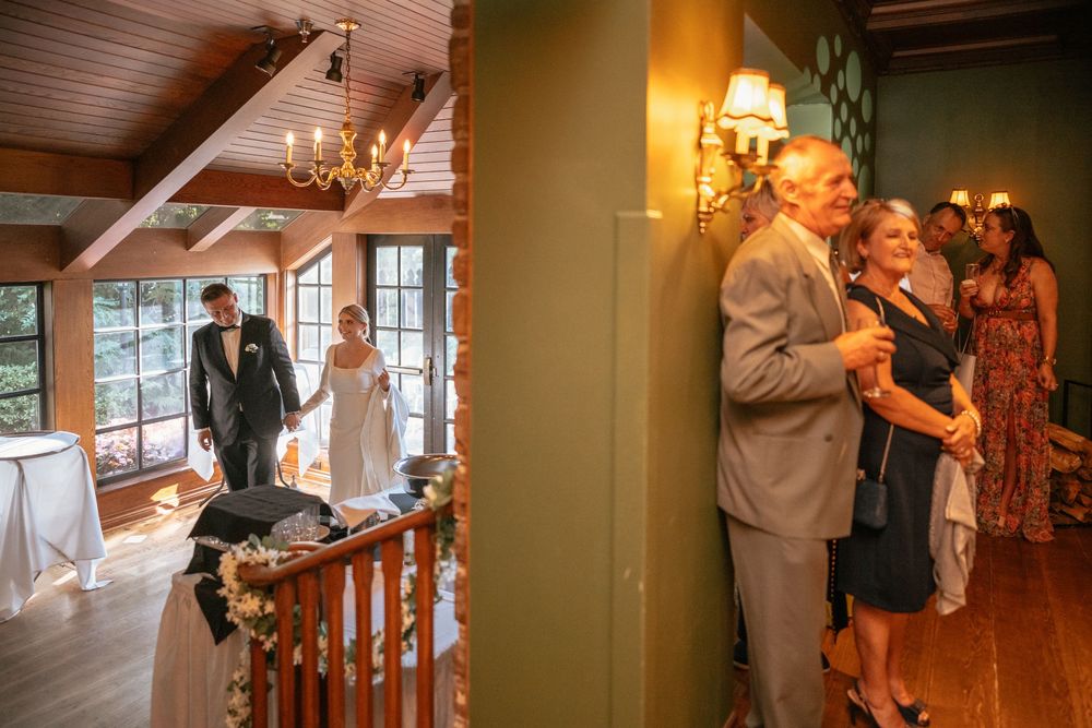 Bride and groom enter wedding venue as guests watch eagerly from nearby hallway.