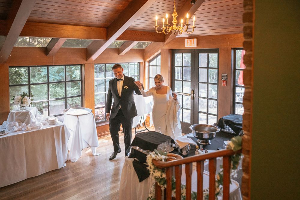 Bride and groom walking hand in hand through elegant wood-beamed wedding venue with chandelier and large windows.
