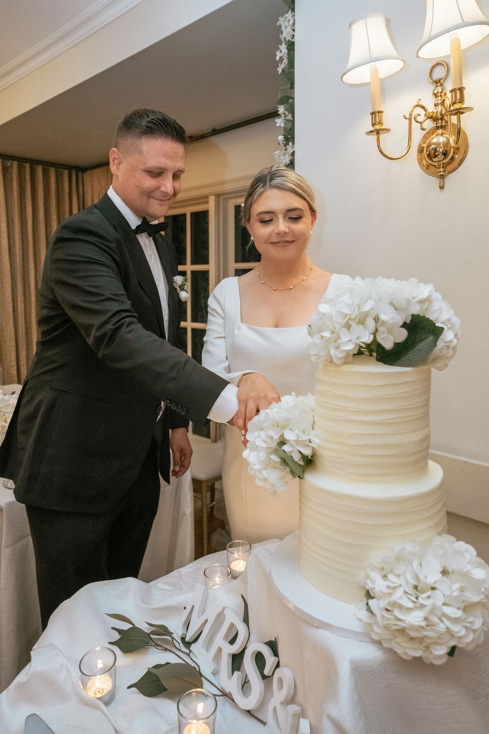 Newlyweds cutting elegant three-tier white wedding cake decorated with hydrangeas.