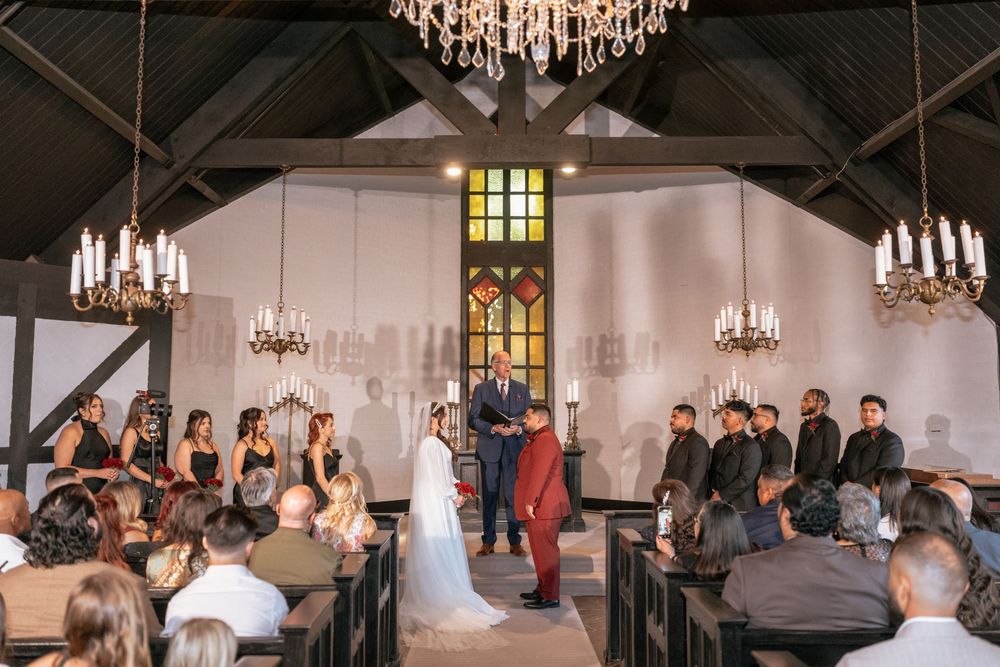 Wedding ceremony in a rustic chapel with chandeliers, bride and groom at altar with wedding party in black and red attire.