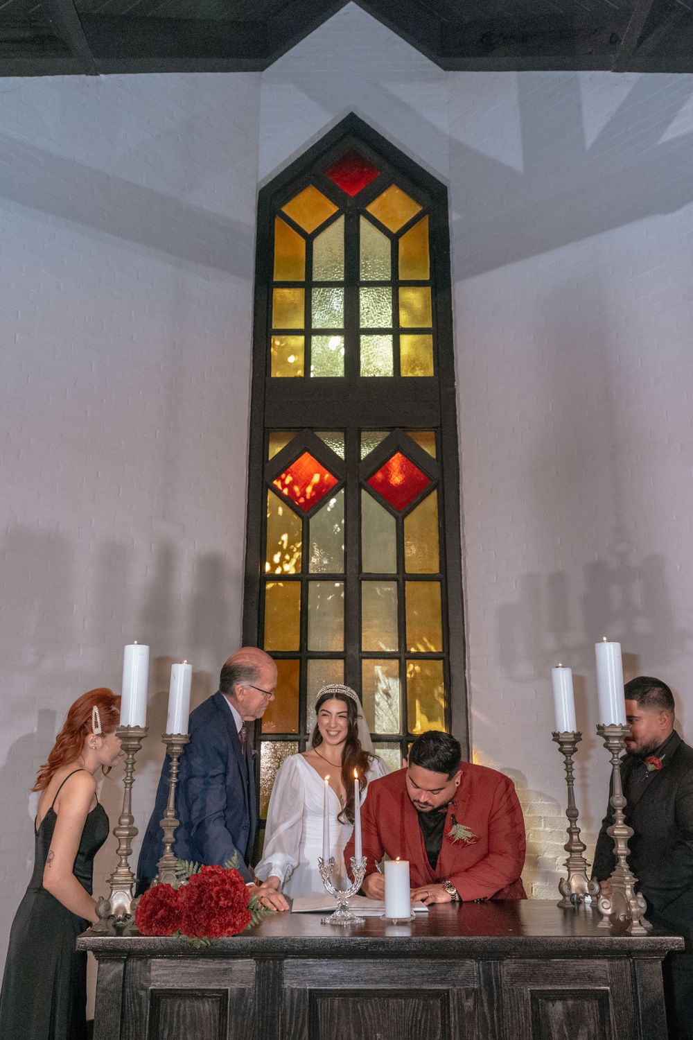 Couple signing wedding documents at a rustic altar with stained glass window and candles in a chapel.
