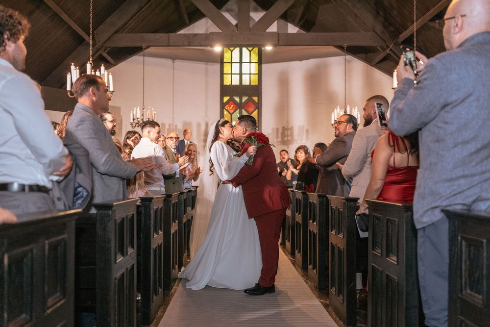 Bride and groom share first kiss at church altar while guests celebrate in wooden pew-lined aisle during wedding ceremony.