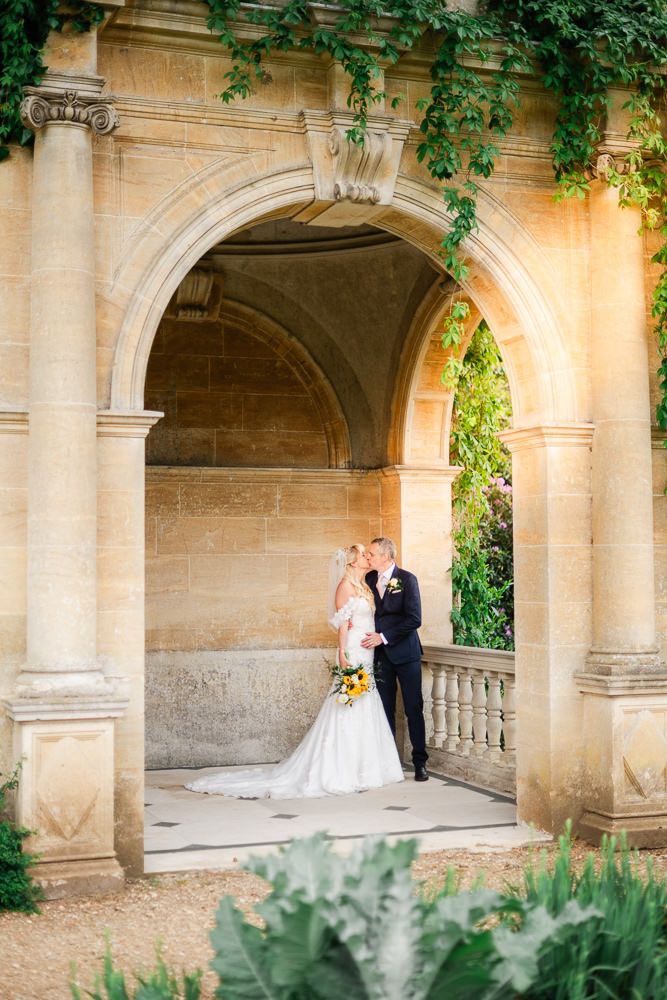 Bride and groom share a romantic kiss under a stone archway adorned with ivy at an elegant outdoor wedding venue.