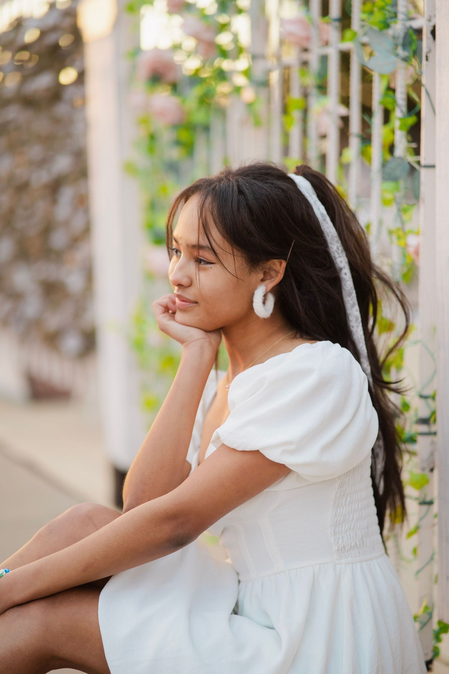 Young woman in white puff-sleeve dress sits thoughtfully beside a vine-covered fence in soft light.