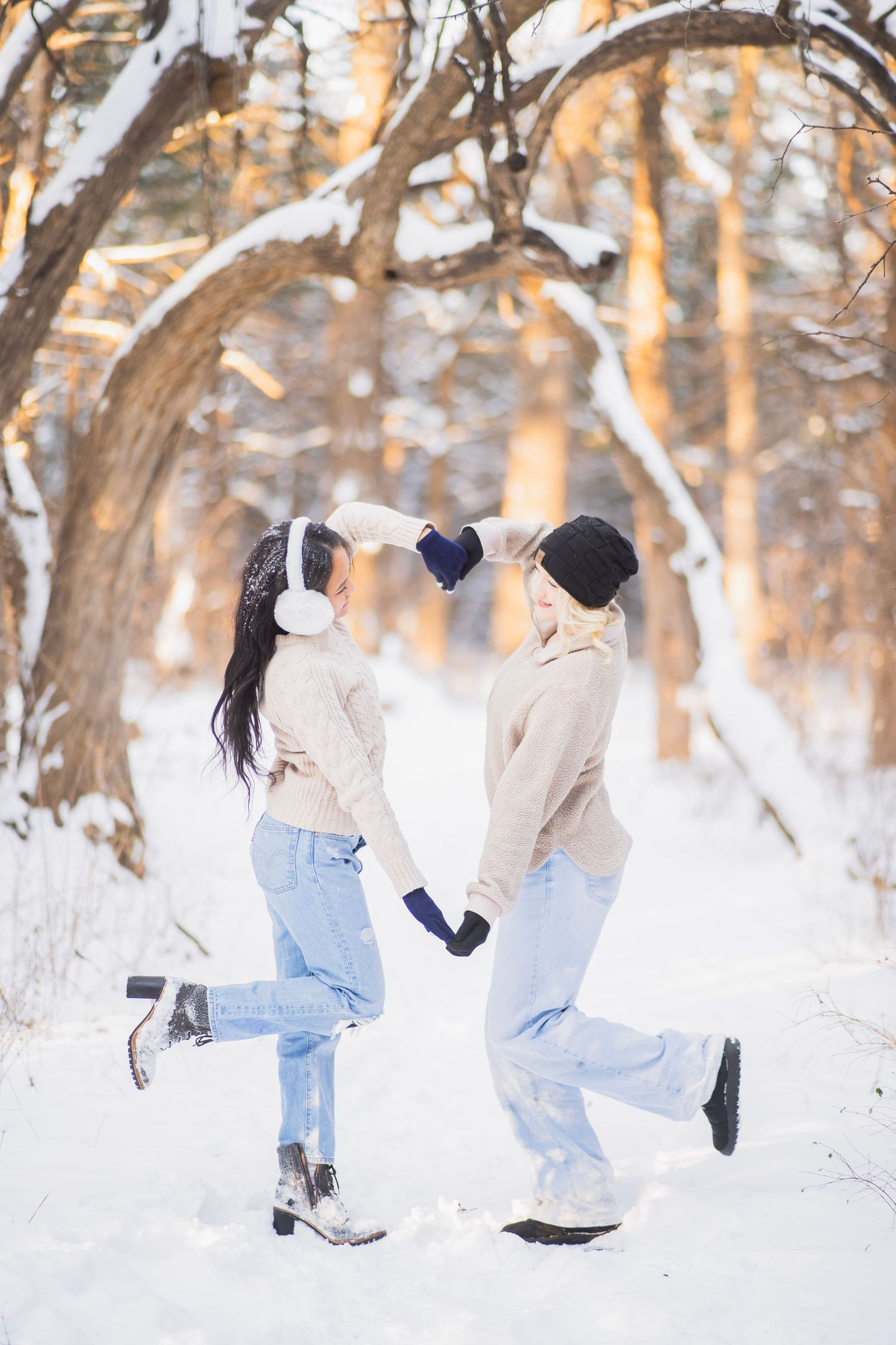 Two young women holding hands and dancing playfully in a snowy winter forest at sunset.